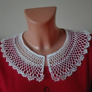 May include: White crocheted collar with a delicate lace pattern. The collar is displayed on a red shirt.