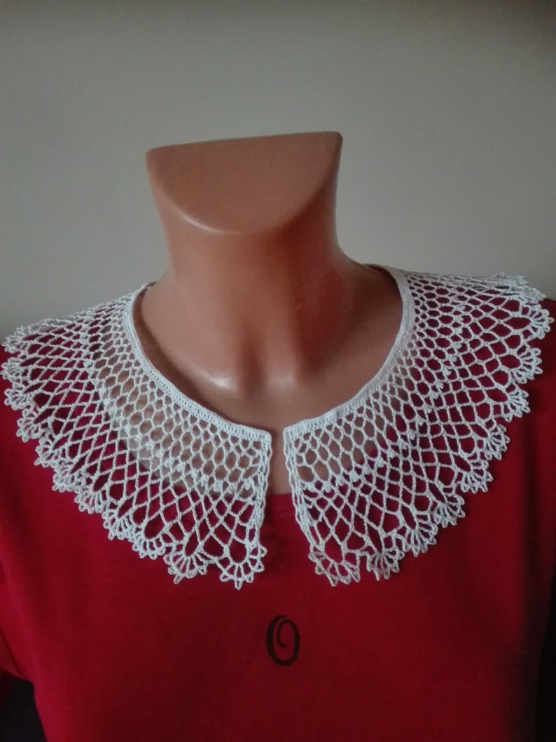 May include: White crocheted collar with a delicate lace pattern. The collar is displayed on a red shirt.