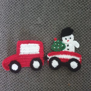 Peut inclure: Une voiture rouge en crochet tirant un wagon avec un bonhomme de neige tenant un sapin de Noël vert en crochet. Le bonhomme de neige porte un chapeau noir.