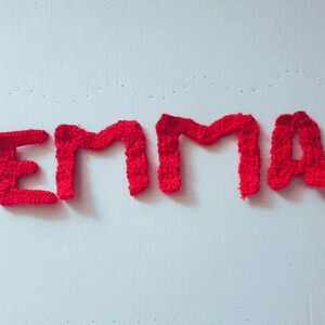 May include: The name "Emma" spelled out in red crocheted letters against a white background.