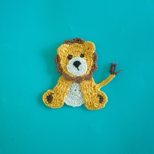 May include: A handmade crochet lion applique in shades of yellow, brown, and cream. The lion has a detailed mane, a cream-colored face, and small black eyes. The applique is set against a solid turquoise background.