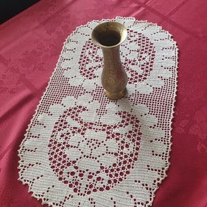 Pode incluir: Um corredor de mesa de crochê de cor creme, em forma oval, com um desenho floral, repousando sobre uma toalha de mesa vermelha. Um pequeno vaso dourado está em cima do corredor. O vaso tem um topo alargado e um padrão detalhado.