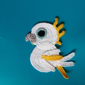 Op de afbeelding: Een witte gehaakte vogel met gele accenten. De vogel heeft een zwart oog en een grijze snavel. De vogel bevindt zich op een blauwe achtergrond.