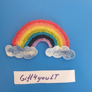 Könnte beinhalten: Ein handgehäkelter Regenbogen mit leuchtenden Farben: Rot, Orange, Gelb, Grün, Blau, Indigo und Violett. Zwei flauschige weiße und blaue Wolken befinden sich am unteren Rand. Der Text "Gift4youLT" steht auf einem weißen Rechteck darunter.
