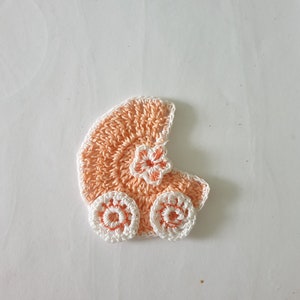 Puede incluir: Una aplicación de cochecito de bebé de ganchillo en color melocotón y blanco. El cochecito tiene dos ruedas y un detalle de flor.