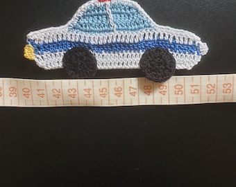 May include: A crocheted police car applique featuring white, light blue, and black yarn with a small red light on the roof and a yellow rear light. A measuring tape is positioned underneath, showing the car spans approximately 4.5 inches in length.