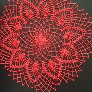Può includere: Un centrino rosso fatto a mano, all'uncinetto con un design traforato dettagliato. Il centrino circolare presenta un motivo ripetuto di motivi floreali e un bordo smerlato. L'intricato pizzo è esposto su uno sfondo scuro.