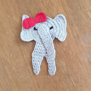 May include: A gray crocheted elephant with a red bow on its head. The elephant is standing on a wooden surface.