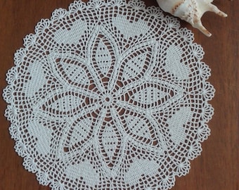 May include: White crocheted doily with a delicate floral pattern, featuring a circular design with a central motif. The doily is on a wooden surface.