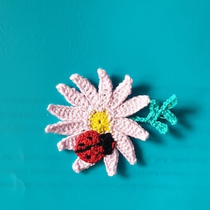 Op de afbeelding: Een roze gehaakte bloem met een gele kern en een rode lieveheersbeestje erop. De bloem is op een turquoise achtergrond.