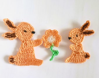 May include: Two crocheted orange bunnies with brown ears and black eyes. One bunny is holding a crocheted orange flower with a white center and green stem.
