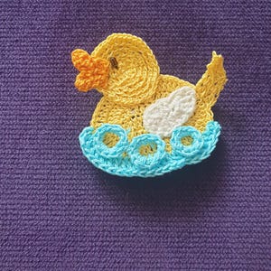 Puede incluir: Un patito hecho a mano con ganchillo en amarillo, naranja, blanco y azul. El patito flota sobre una base azul con forma de agua. El patito tiene un cuerpo amarillo, un pico naranja y un ala blanca. El fondo es de color morado liso.