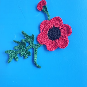 Puede incluir: Una flor de amapola de ganchillo hecha a mano con pétalos rojos y un centro negro. La flor tiene un tallo y hojas verdes. La amapola está sobre un fondo azul brillante.