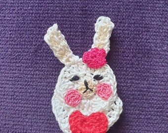 May include: A handmade crochet bunny applique with a white body, pink cheeks, and a red heart. The bunny has a pink flower on its head and black eyes. The applique is likely for crafting or embellishment.