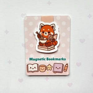 May include: A magnetic bookmark featuring a cartoon red panda eating noodles and reading a book. The bookmark has a pink background with white polka dots and the words "Magnetic Bookmarks" at the bottom.