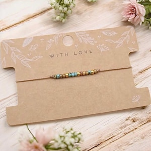Könnte beinhalten: Ein Armband mit Türkis- und goldfarbenen Perlen liegt auf einer braunen Karte mit der Aufschrift "WITH LOVE". Die Karte hat ein florales Design und liegt auf einer Holzoberfläche mit Blumen.
