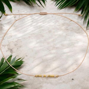 Könnte beinhalten: Zarte Halskette mit einem dünnen, beigefarbenen Band und einer Reihe kleiner gold- und silberfarbener Perlen. Die Halskette hat einen Schiebeknotenverschluss. Der Hintergrund ist eine strukturierte Oberfläche mit grünen Blättern.