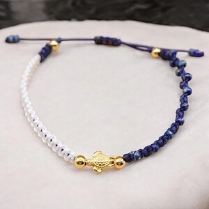 Könnte beinhalten: Ein blaues und weißes Perlenarmband mit einem goldenen Schildkrötenanhänger. Das Armband ist verstellbar und hat einen verschiebbaren Knotenverschluss.