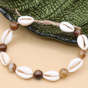 Könnte beinhalten: Ein weißes und braunes Muschel-Armband mit einer braunen Kordel. Das Armband besteht aus kleinen, weißen Muscheln und braunen Perlen.