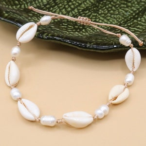Könnte beinhalten: Ein weißes Muschel- und Perlenarmband mit einer braunen Schnur. Das Armband ist mit einem Knoten gebunden und hat ein schlichtes Design.