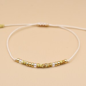 Könnte beinhalten: Ein weißes Armband aus Schnur mit goldenen und grünen Perlen. Das Armband ist verstellbar und kann am Handgelenk oder am Knöchel getragen werden.
