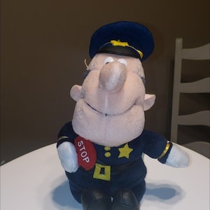 May include: A plush toy police officer with a blue uniform, a yellow star, and a red stop sign. The toy has a white face with a large nose and a friendly smile.