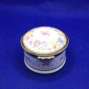 May include: A small, round, white porcelain trinket box with a floral design on the lid. The lid has a gold trim and the box has a gold band around the middle.