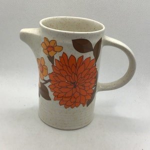 May include: A white ceramic pitcher with a speckled glaze and a floral design in shades of orange, yellow, and brown. The pitcher has a handle and a spout.