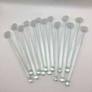 May include: A collection of thirteen clear glass stirrers, each featuring a long, slender stem and a decorative, textured floral-shaped top. These transparent beverage stirrers are arranged in a fan-like formation against a plain background.