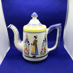 May include: A white ceramic teapot with a blue and yellow floral design and a woman in a yellow dress holding a flower. The teapot has a blue handle and a blue spout.