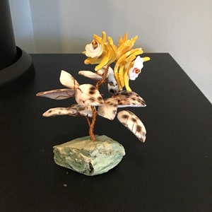 May include: A small, decorative tree made of seashells and wire. The tree has white and yellow seashells for flowers and brown and white seashells for leaves. The tree is sitting on a green rock.
