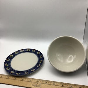 K&ouml;nnte beinhalten: Ein ovaler Keramikteller mit blauem Rand und floralem Design, neben einer wei&szlig;en Keramikschale. Der Teller hat eine wei&szlig;e Mitte. Die Sch&uuml;ssel ist rund und hat eine glatte, cremefarbene Oberfl&auml;che. Ein Holzlineal befindet sich im Vordergrund.
