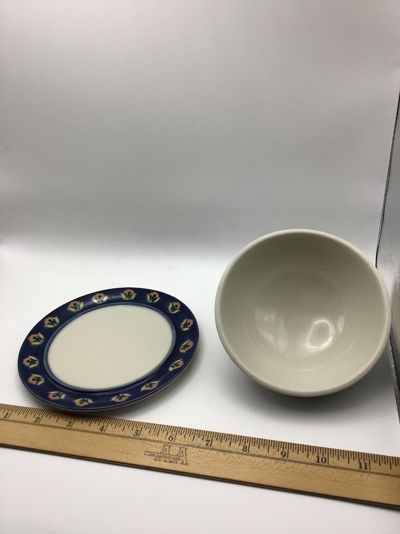 K&ouml;nnte beinhalten: Ein ovaler Keramikteller mit blauem Rand und floralem Design, neben einer wei&szlig;en Keramikschale. Der Teller hat eine wei&szlig;e Mitte. Die Sch&uuml;ssel ist rund und hat eine glatte, cremefarbene Oberfl&auml;che. Ein Holzlineal befindet sich im Vordergrund.