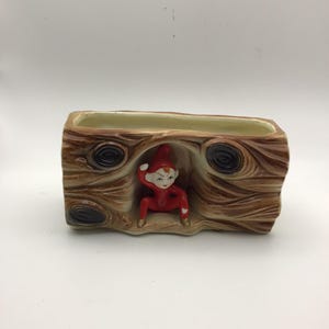 May include: A rectangular ceramic planter shaped like a brown log with a textured wood grain pattern and dark knots. A small, red-skinned figure with a pointed hat and white facial features is positioned in a hollow opening in the center.