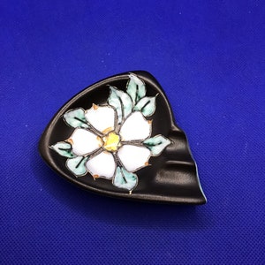 Puede incluir: Un cenicero de cerámica negra con un diseño de flor blanca. La flor tiene hojas verdes y un centro amarillo.