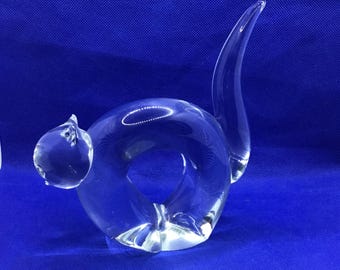May include: A clear glass sculpture in the shape of a snail. The snail has a rounded body with a large opening, a small head, and a curved tail. The sculpture is on a blue background.