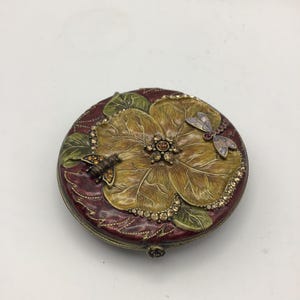May include: A round, decorative trinket box with a floral design. The box features a large, yellow flower with a jeweled center, surrounded by green leaves and a burgundy background. A bee and dragonfly are also featured.