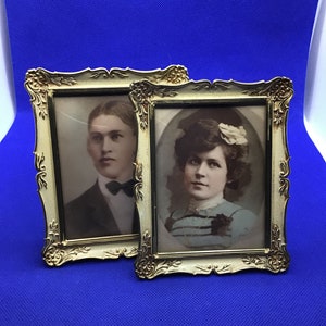May include: Two ornate gold-colored picture frames with glass fronts, each holding a black and white portrait of a person. The frame on the left features a man with a bow tie, while the frame on the right features a woman with a flower in her hair.