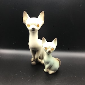 May include: Two ceramic cat figurines. One is tall and seated, the other is smaller and sitting. Both cats are white with tan accents and have orange rhinestone eyes. The background is black.