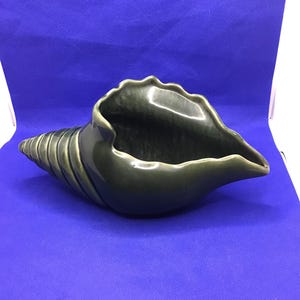 May include: A dark green ceramic seashell-shaped decorative bowl. The bowl has a spiral design and a wavy rim. The interior is a darker shade of green. The bowl is on a blue background.