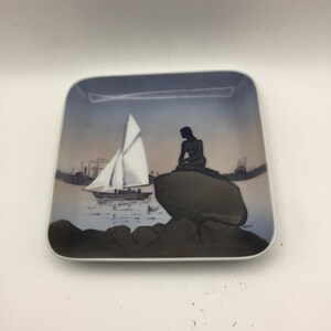 May include: Square ceramic plate with a nautical scene. The plate features a sailboat with white sails, a dark silhouette of a person sitting on a rock, and a blue and brown background.