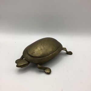 May include: A brass turtle figurine with a smooth, rounded shell and four distinct legs. The metal has a muted, aged gold-toned finish with visible surface texture and natural patina, showing a simple, stylized form of a turtle in a stationary position.