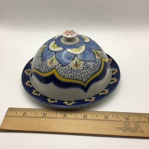 K&ouml;nnte beinhalten: Eine Keramik-Butterdose mit gew&ouml;lbtem Deckel und passender Platte. Die Dose hat ein blaues Blumenmuster mit gelben und orangefarbenen Akzenten. Der Deckel hat einen kleinen Knopf oben. Der Durchmesser von Platte und Deckel betr&auml;gt ca. 15 cm.