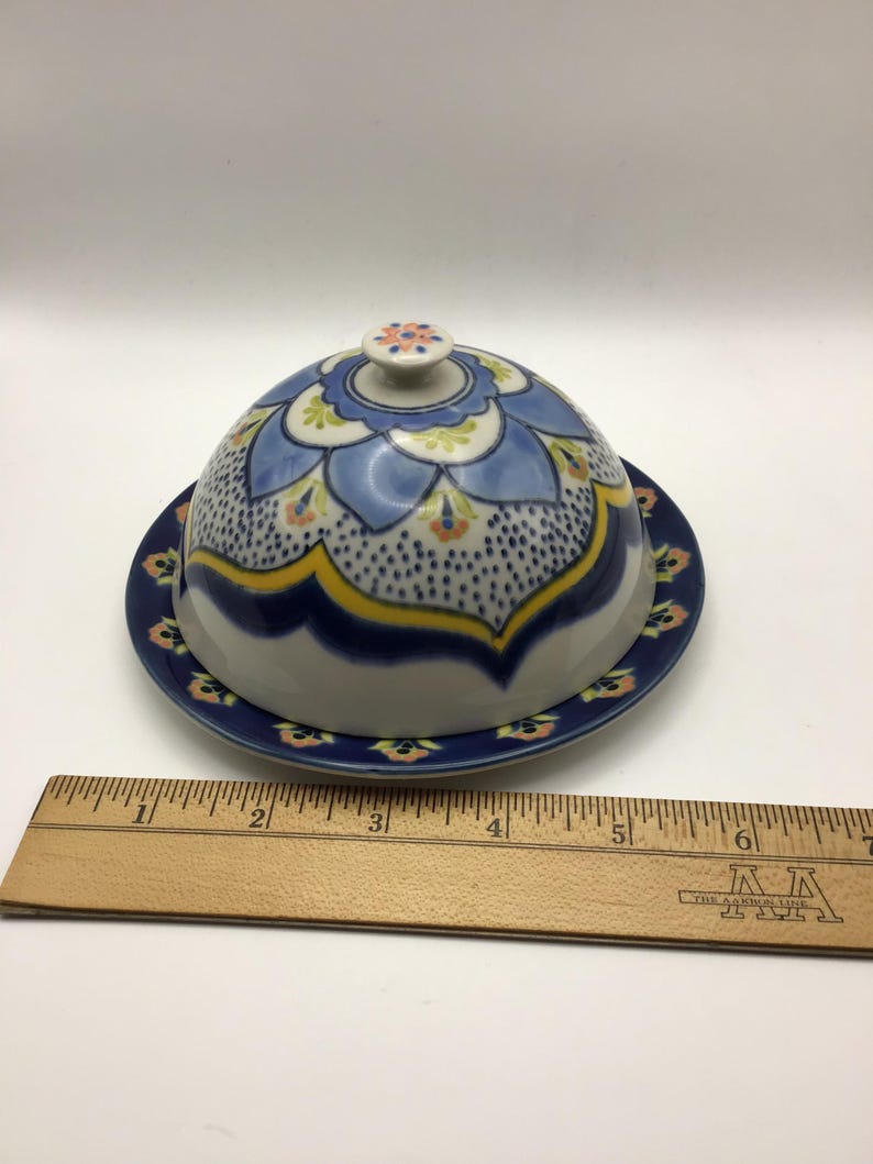 K&ouml;nnte beinhalten: Eine Keramik-Butterdose mit gew&ouml;lbtem Deckel und passender Platte. Die Dose hat ein blaues Blumenmuster mit gelben und orangefarbenen Akzenten. Der Deckel hat einen kleinen Knopf oben. Der Durchmesser von Platte und Deckel betr&auml;gt ca. 15 cm.