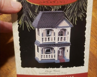 May include: A white box with a red border and the text "Keepsake Ornament" on the front. The box contains a small, white, two-story house ornament with a black roof and red door. The text "Cozy Home" and "Nostalgic Houses and Shops" is printed on the front of the box. The text "Collector's Series" is printed in red on the bottom of the box.