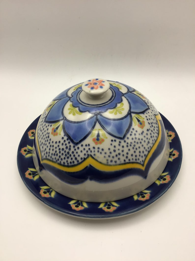 K&ouml;nnte beinhalten: Eine Keramik-Butterdose mit gew&ouml;lbtem Deckel und passendem Teller. Der Deckel hat ein blaues Blumenmuster mit gelben und orangefarbenen Akzenten. Teller und Deckel haben einen dunkelblauen Rand mit floralen Mustern.