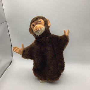 May include: A plush monkey hand puppet with dark brown fur, a light tan face, and white chin fur. The puppet features two outstretched arms with tan hands, dark eyes, and a neutral expression, designed to be worn on the hand for performance.