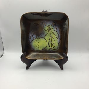 May include: Square ceramic dish with a brown and gold glaze. The dish features a green and blue fruit design. The dish is displayed on a dark brown wooden stand. The dish is a decorative item.