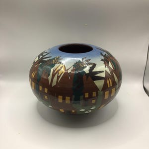 May include: A round ceramic vase with a wide opening and a dark interior. The exterior features a landscape design with trees, buildings, and geometric shapes in brown, green, and yellow against a blue sky.