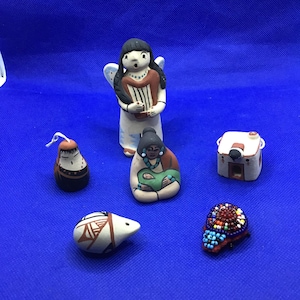 May include: A collection of small, handcrafted ceramic figurines. The figurines include an angel, a woman holding a baby, a small house, a hedgehog, a beaded object, and a small figure with a hat. The figurines are painted with various colors and designs.